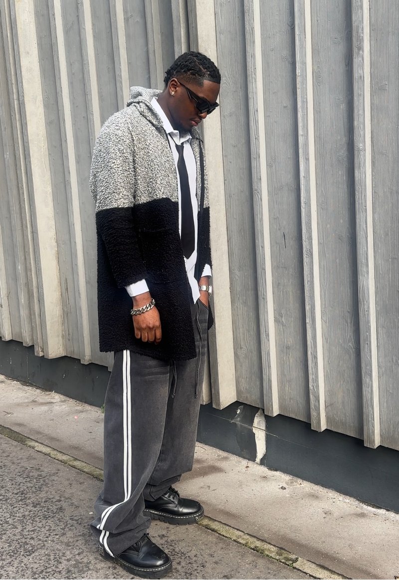 Homme portant des lunettes de soleil, manteau à capuche noir et gris, chemise blanche avec cravate noire, pantalon gris à rayures blanches et chaussures noires, debout près d'un mur gris.