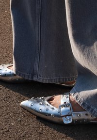 Sølvmetallic flate sko med spiss tå, naglede detaljer og en spenne. Kombinert med vide denimbukser i en lys blå farge.