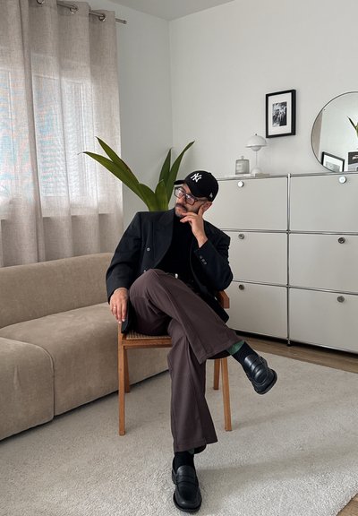 Blazer negro sobre un cuello de tortuga negro, pantalones marrones, usando una gorra negra. Sentado en una silla de madera en una habitación de tonos neutros con una planta y un espejo.