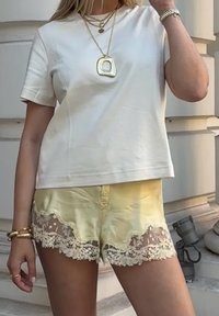 Camiseta blanca de manga corta combinada con pantalones cortos de satén amarillos con borde de encaje y detalle de botón. Los accesorios incluyen collares en capas y pulseras.