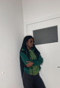 Sudadera con cremallera verde y negra de manga larga, llevada con pantalones negros. La persona tiene el cabello largo trenzado y está situada contra una pared blanca.