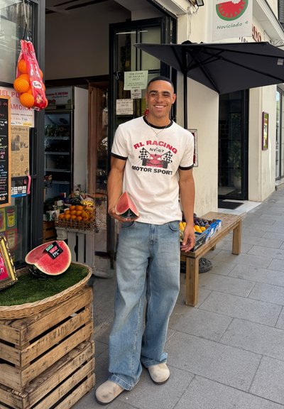 Una rodaja de sandía sostenida por una persona con una camiseta blanca con gráficos rojos, pantalones vaqueros anchos de color azul claro y calzado beige, frente a una tienda de frutas.
