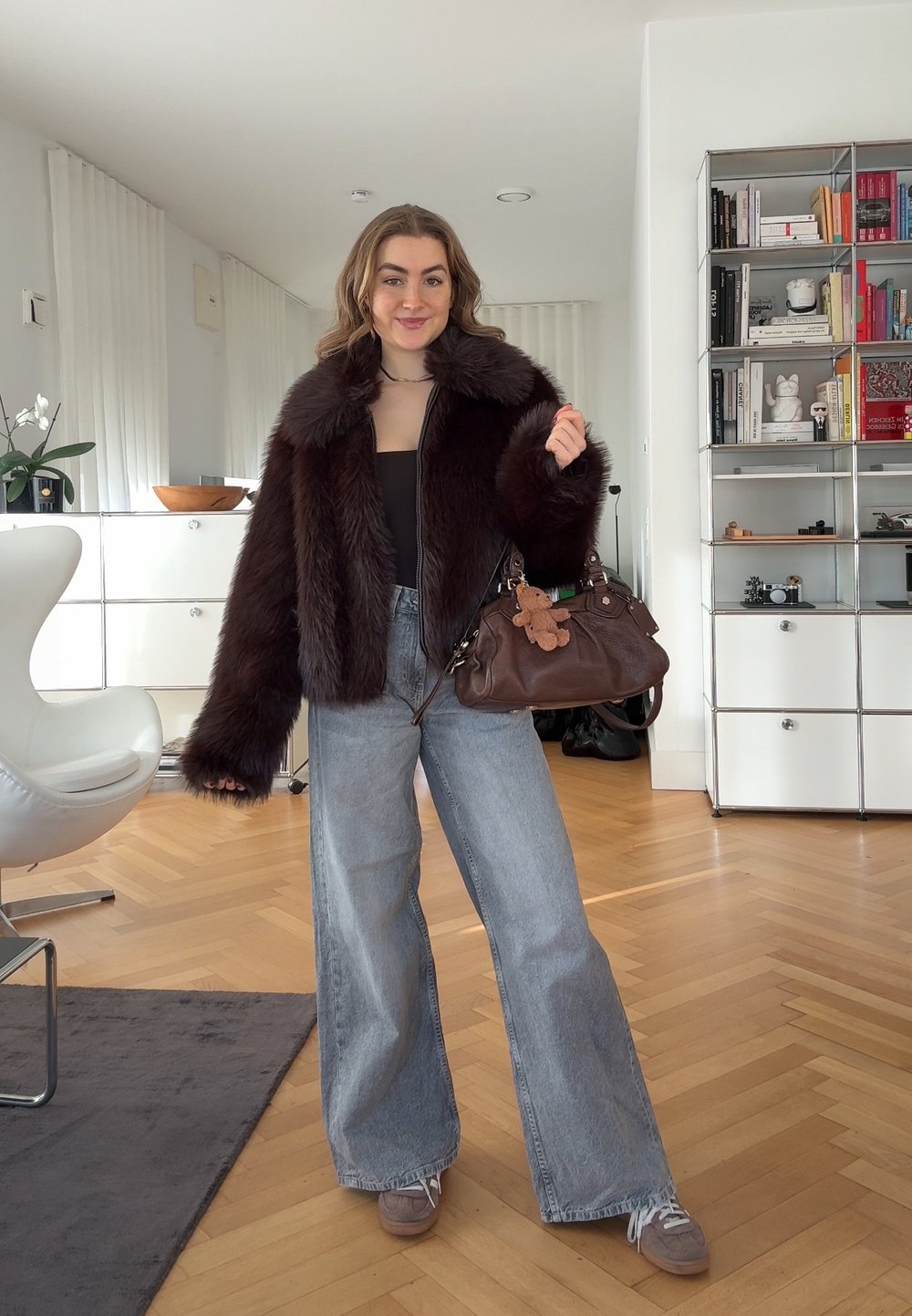 Jeune femme en jean à jambes larges et veste en fourrure sombre tenant un sac à main marron avec un porte-clés en forme d'ourson dans un salon moderne.