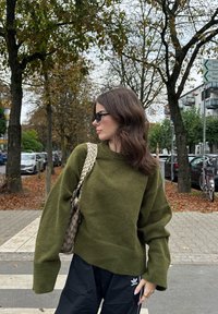Maglione oversize verde oliva con orlo a coste, abbinato a pantaloni jogger neri e una borsa beige con motivo. La scena mostra foglie autunnali.