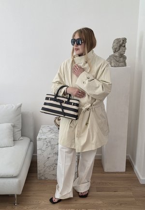 Mujer con abrigo tipo gabardina color marfil, pantalones anchos blancos, gafas de sol negras sosteniendo un bolso de mano a rayas blancas y negras en un ambiente interior moderno con busto escultórico.