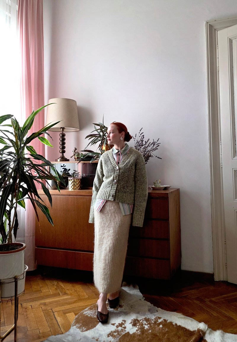 Knitted green cardigan with large buttons worn over a striped shirt and a long, fluffy beige skirt. Wooden furniture and indoor plants in the background.