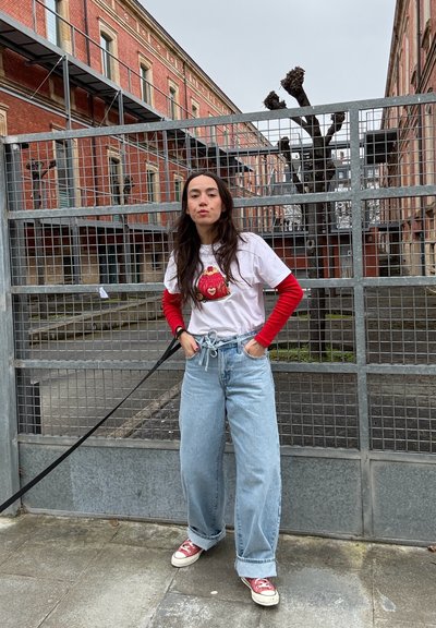 Camiseta blanca con mangas largas rojas, con diseño gráfico, combinada con vaqueros de pierna ancha de color azul claro y zapatillas rojas, frente a una cerca de metal.