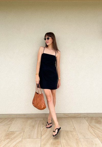 Vestido negro ajustado con tirantes finos, acompañado de un bolso de mano marrón trenzado. Chanclas negras y pelo lacio completan el look. Fondo neutro.