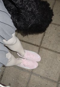 Ružové tenisky s bielymi akcentmi, nosené s bielymi pančuchami a šedou plisovanou sukňou, čiastočne zakryté čiernou kožušinovou textúrou.