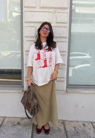 Mujer con cabello largo y oscuro, con gafas, camiseta blanca con estampado de botas rojas, falda beige, zapatos burdeos, sosteniendo bolso marrón, de pie al aire libre.