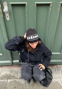 Juodas vilnos paltas su apvartu, derinamas su laisvais pilkais kelnėmis, juoda beanie kepurė su "obey" logotipu ir smailiomis juodomis aukštakulnėmis botais.