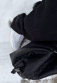 Hvit joggesko på snø, kombinert med svarte bukser i teksturert stoff og en svart skinnveske med glidelås og håndtak. Snødetaljer er synlige.