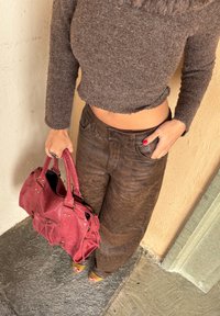 A woman wearing a brown long-sleeved cropped sweater and brown trousers stands with one hand in her pocket, holding a red handbag in the other.