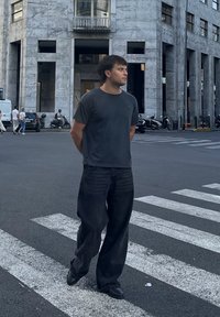 Čierne tričko, voľné čierne denimové nohavice a čierne topánky. Stojí na prechode pre chodcov, s mramorovou budovou a mestským prostredím v pozadí.