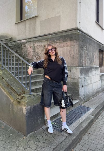 Chaqueta negra con cremallera y rayas blancas, pantalones cortos de mezclilla hasta la rodilla, calcetines blancos y zapatos negros. Bolso negro con un encanto blanco.