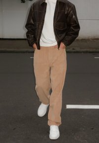 Chaqueta de cuero marrón, sudadera blanca, pantalones de pana beige y zapatillas blancas. El atuendo presenta un corte relajado con materiales texturizados.