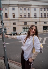 Camisa blanca abotonada con mangas de encaje, pantalones deportivos marrones con rayas laterales y un bolso amarillo con un detalle de felpa. Fondo urbano.