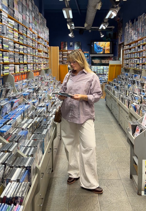 Una persona si trova in una corsia di un negozio, esaminando un DVD. Indossa una camicia a righe viola chiaro, pantaloni bianchi larghi e scarpe marroni.