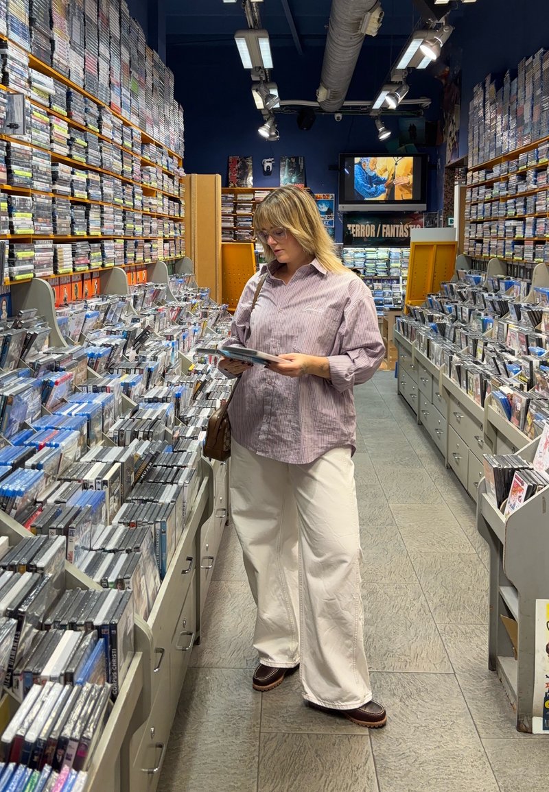 Una persona si trova in una corsia di un negozio, esaminando un DVD. Indossa una camicia a righe viola chiaro, pantaloni bianchi larghi e scarpe marroni.