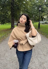 Kvinna i beige kappa och jeans håller en iskall kaffekopp och en ljusbeige handväska medan hon går längs en parkstig med träd och bänkar.