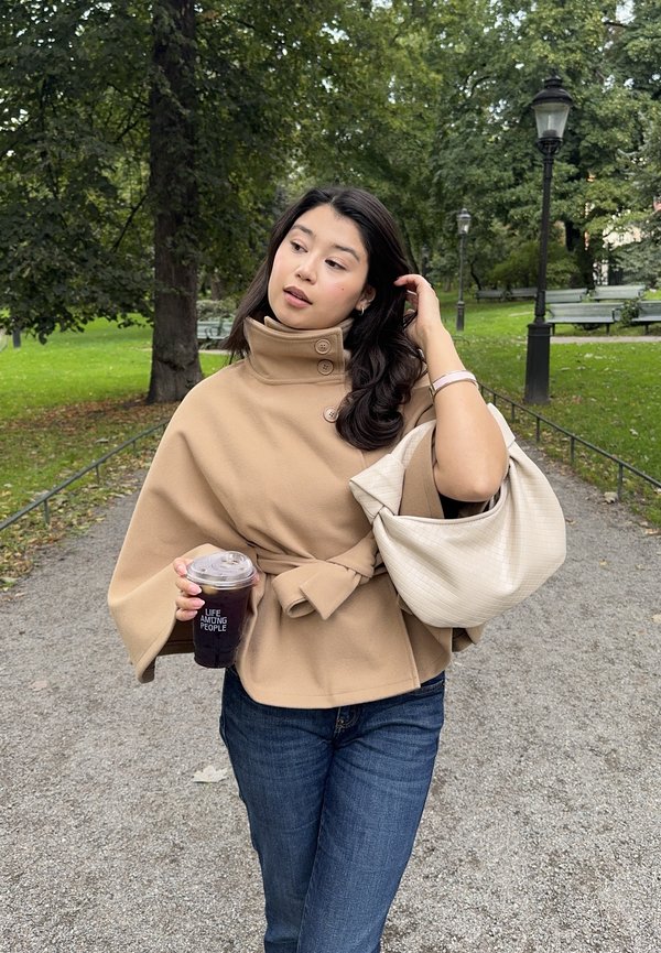 Mujer con abrigo beis y vaqueros sostiene una taza de café helado y un bolso beis claro mientras camina por un sendero del parque rodeado de árboles y bancos.