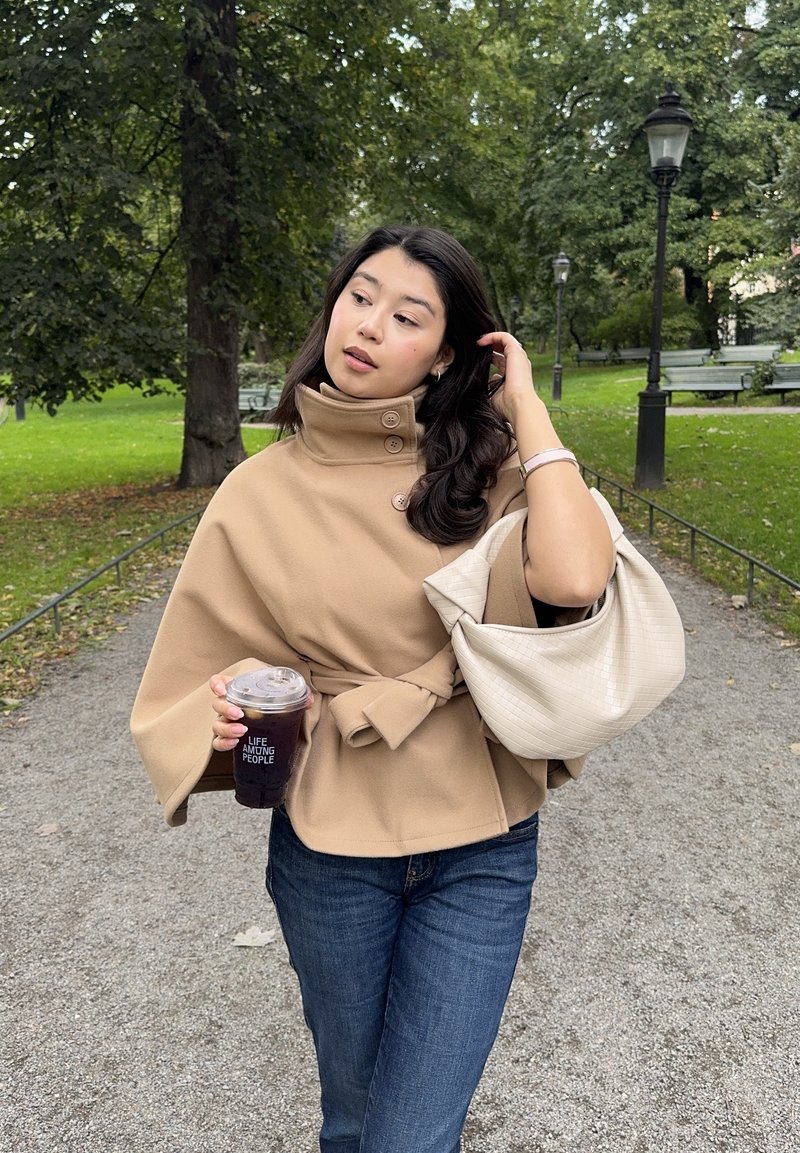 Frau in beigem Cape-Jackett und Jeans hält einen Eiskaffee und eine hellbeige Handtasche, während sie den Parkweg mit Bäumen und Bänken entlanggeht.