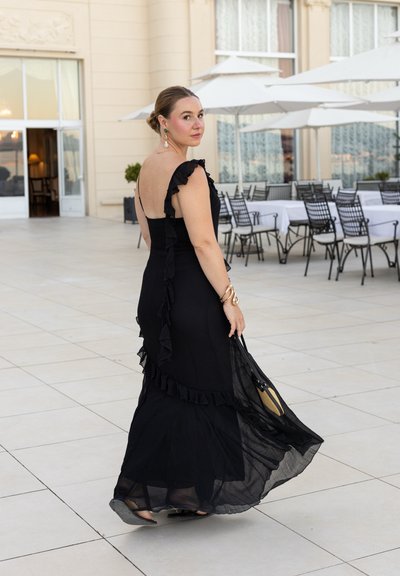 Vestido negro con acentos fruncidos y un corsé ajustado, que fluye hacia una falda transparente de capas. El tejido parece ligero y texturizado.