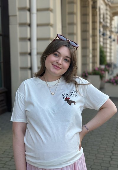 Camiseta blanca de manga corta con un gráfico de un perro usando un sombrero rojo, acompañado del texto "MAISON PEPITA." La persona también lleva collares en capas.