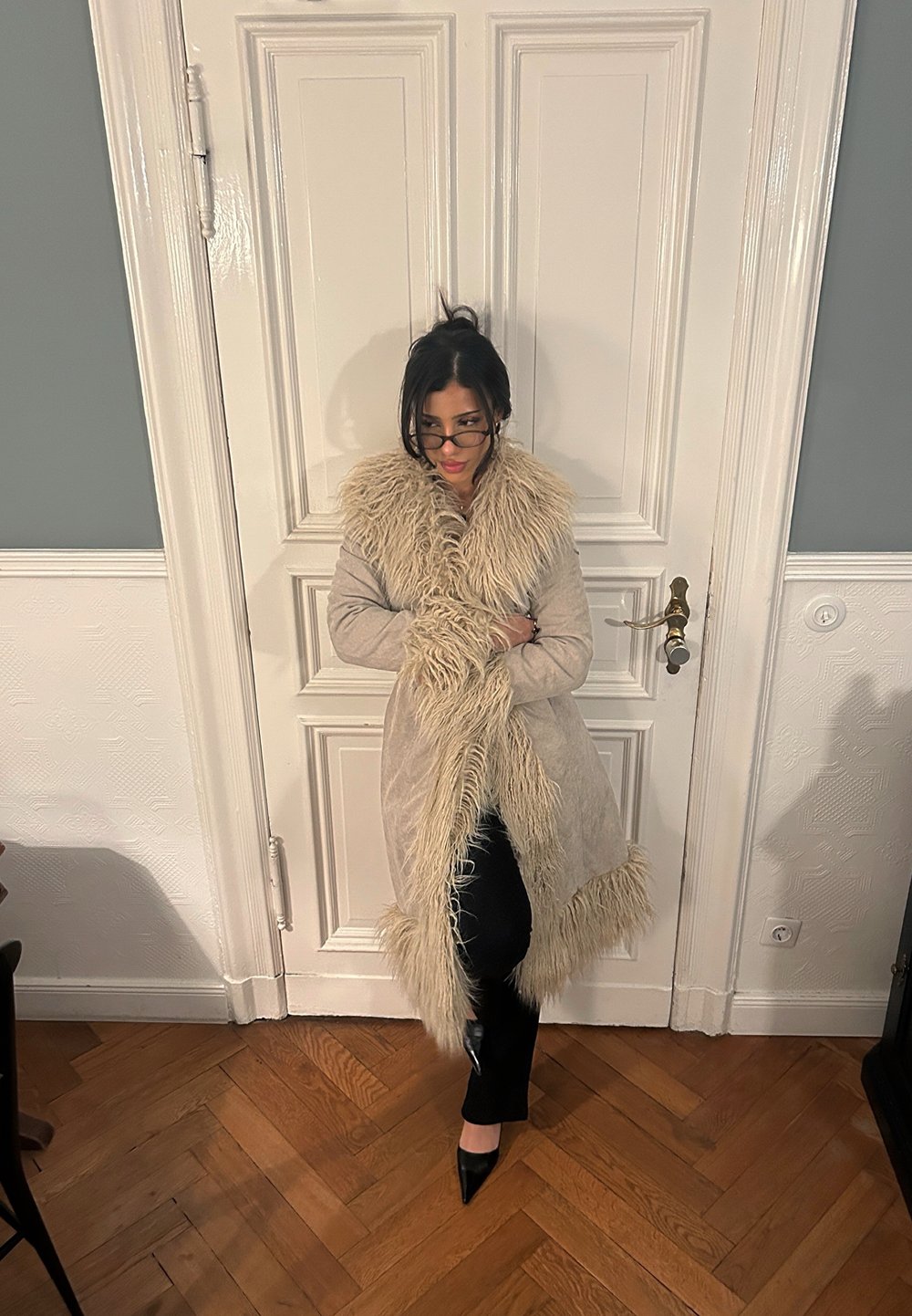 Manteau long beige avec col et poignets en fourrure, dotés d'une finition texturée, accompagné de pantalon noir et de talons pointus noirs sur un sol en bois.