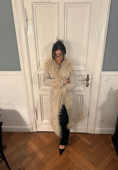 Abrigo largo beige con cuello y puños de piel, acabado texturizado, combinado con pantalones negros y tacones puntiagudos negros sobre un suelo de madera.