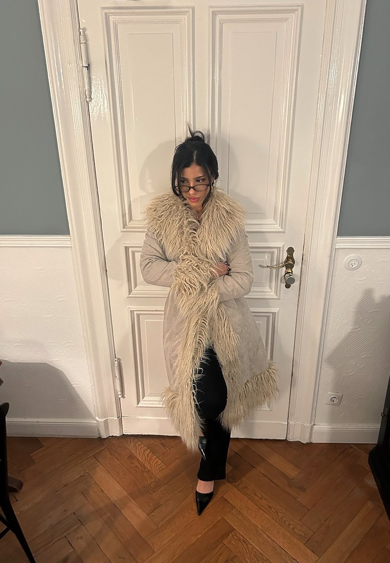 Manteau long beige avec col et poignets en fourrure, dotés d'une finition texturée, accompagné de pantalon noir et de talons pointus noirs sur un sol en bois.