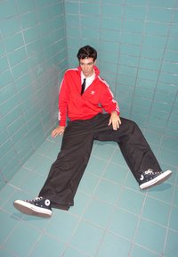 Rode Adidas trainingsjack met witte strepen, zwarte wijde broek, zwarte Converse sneakers. Zittend in een betegelde hoek met turquoise muren.