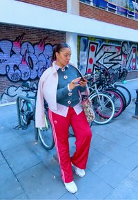 Røde joggebukser med hvite striper, grå genser med blomsterdetaljer, rosa kort jakke, og mønstrete veske; person som bruker en smarttelefon.