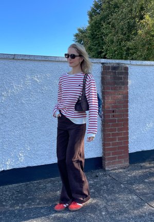 Camisa de rayas rojas y blancas de manga larga, pantalones negros de pierna ancha, zapatos rojos con rayas blancas. Pequeño bolso negro, se apoya contra una pared de ladrillos.