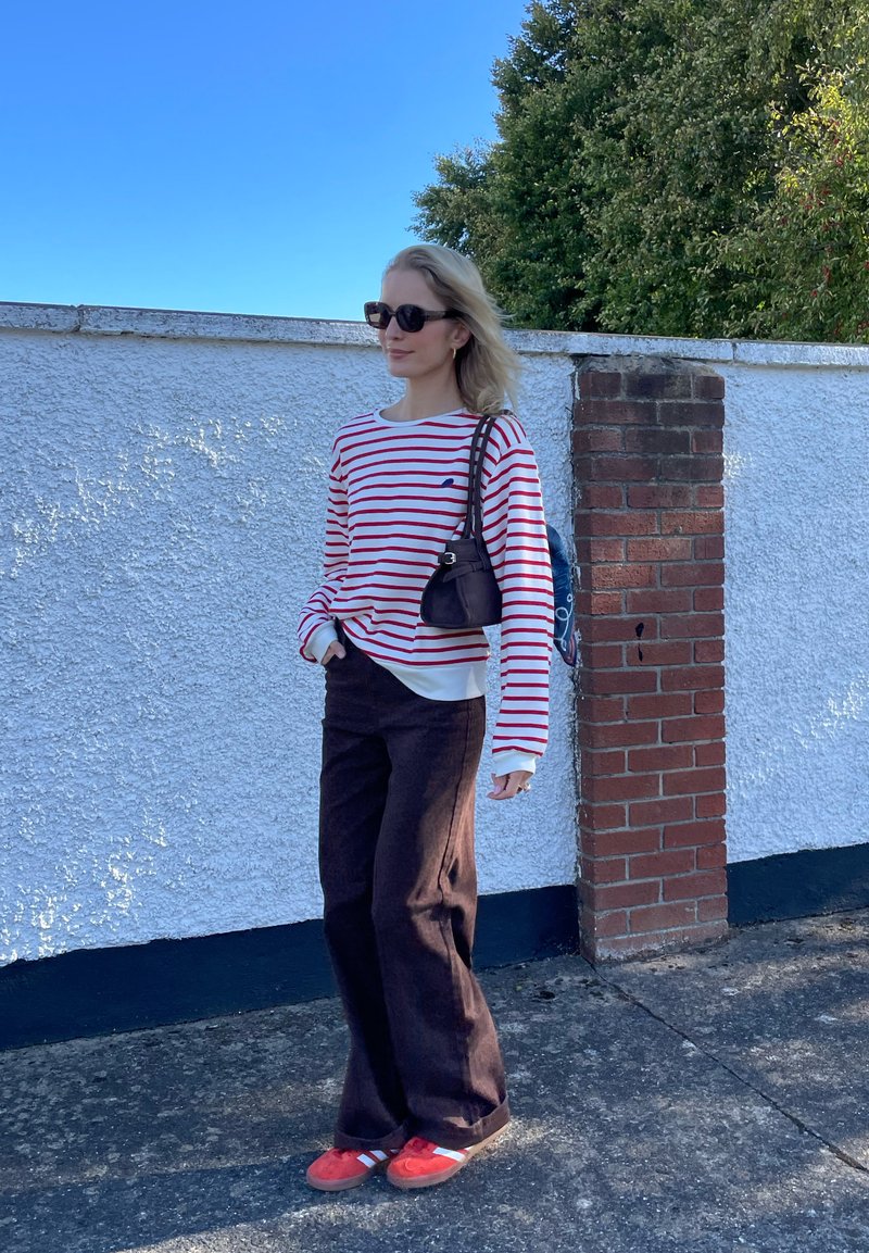 Camicia a righe rossa e bianca a maniche lunghe, pantaloni neri a gamba larga, scarpe rosse con strisce bianche. Piccola borsa nera, si appoggia contro un muro di mattoni.