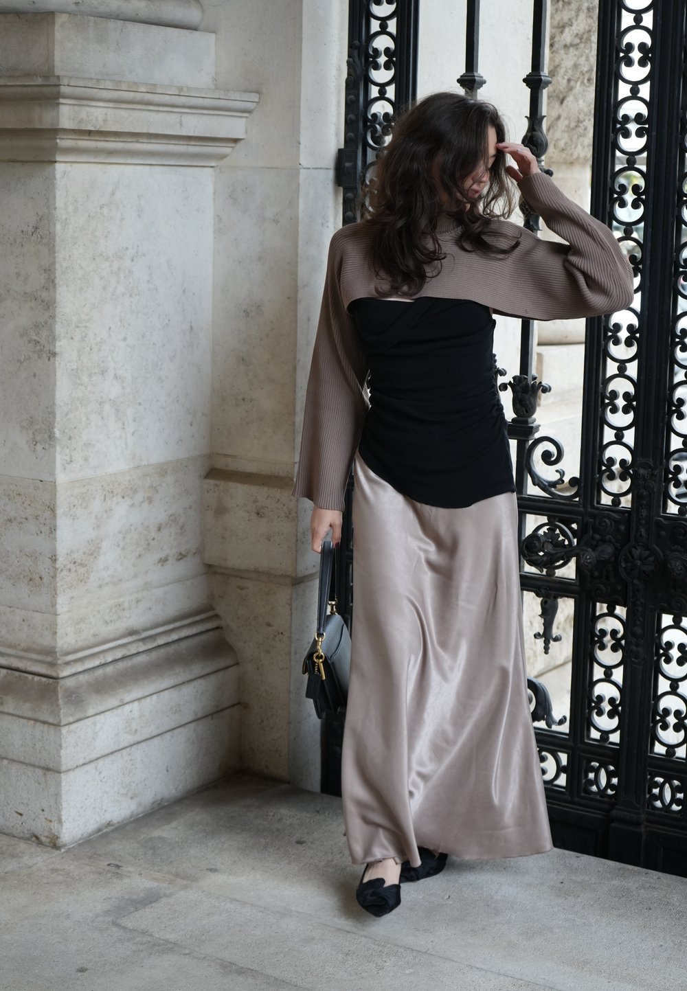 Top aderente nero, sovrapposizione beige a maniche lunghe a coste e gonna lunga in satin beige. Scarpe flat nere e una borsa a tracolla piccola, in piedi vicino a cancelli ornati.