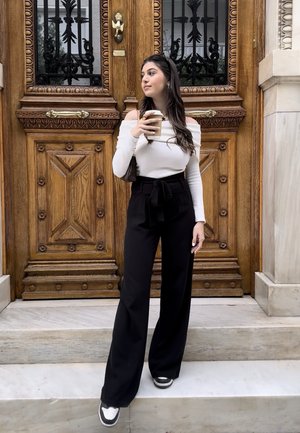 Joven mujer con pantalones negros y top blanco sin hombros, de pie sobre escalones de mármol, sosteniendo una taza de café frente a una puerta doble de madera.