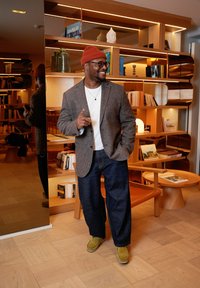 Un homme portant un blazer marron à motifs, une chemise blanche et un jean foncé, assortis de chaussures vertes et d'un bonnet orange, se tient dans une pièce moderne.