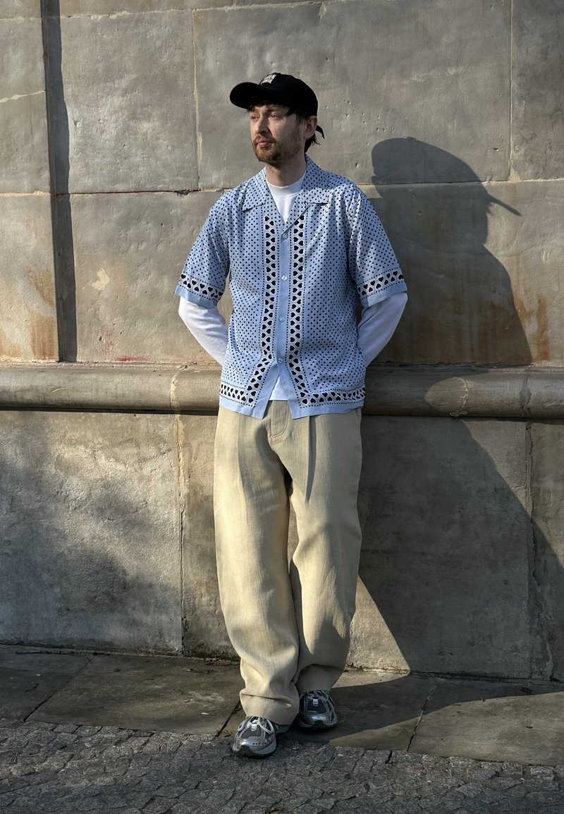 Homme en pantalon beige, chemise blanche à manches longues sous une chemise bleue à manches courtes à motifs, casquette noire et baskets, debout contre un mur en pierre.