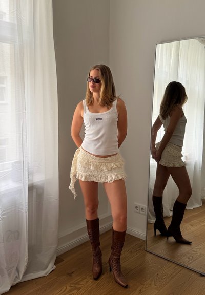 Top de tirantes blanco con textura acanalada y logo negro, falda corta de encaje con varias capas, y botas de tacón altas marrones. El espejo refleja el modelo y su entorno.