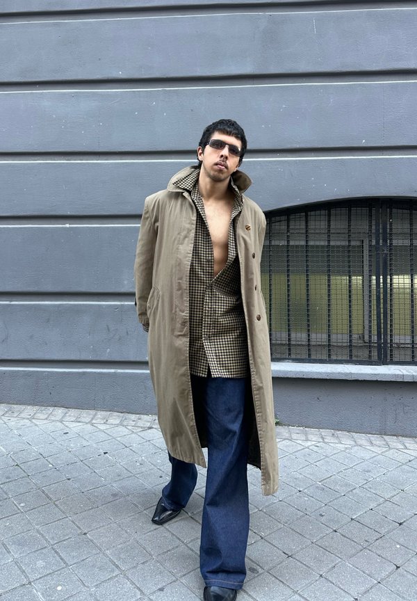 Mand iført beige trenchcoat, åben ternet skjorte, mørke jeans og solbriller, stående på byens fortov mod en grå væg med buet vindue.