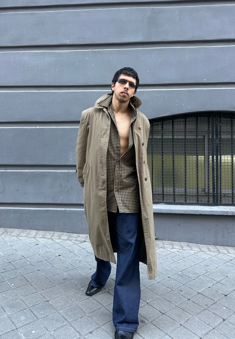 Mann trägt beigefarbenen Trenchcoat, offenes kariertes Hemd, dunkle Jeans und Sonnenbrille, steht am Bürgersteig der Stadt vor einer grauen Wand mit bogenförmigem Fenster.