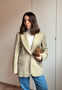 Femme portant un blazer beige, un col roulé blanc, un jean bleu, tenant une pochette marron tissée, debout à l'intérieur contre un mur texturé.