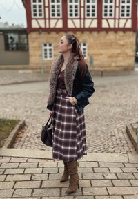 Jupe à carreaux marron avec une fente latérale, associée à une veste foncée avec un col en fourrure, portant des bottes à talons hauts couleur beige, tenant un sac à main.