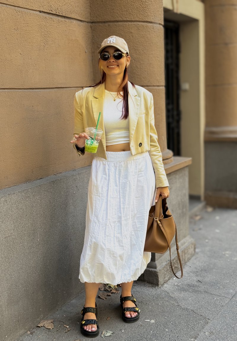 Blazer jaune sur un haut blanc court et une jupe blanche fluide. Les accessoires comprennent une casquette beige, des lunettes de soleil rondes et un sac brun.