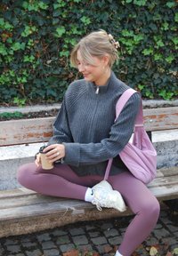 Grijze geribbelde trui met een halve rits, roze rugzak, hooggepolsterde mauve legging en witte sneakers, terwijl ze zittend een koffiekopje vasthoudt.