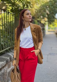 Sacou bej, tricou alb, pantaloni roșii cu talie înaltă; ținând o geantă maro; mediu exterior cu vegetație în fundal, lumină naturală.