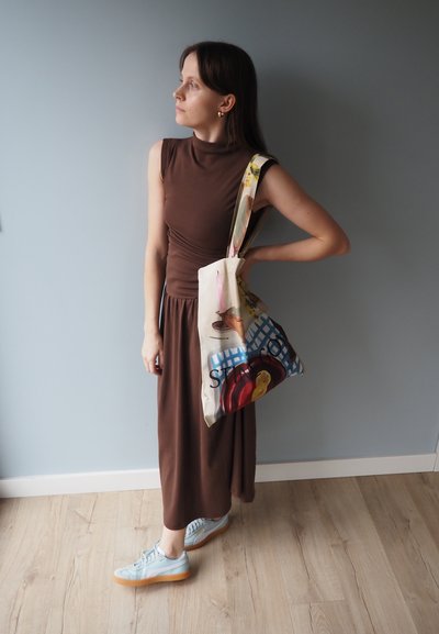 Top sin mangas marrón ajustado y falda larga, zapatillas azules, sosteniendo un bolso de crema con un diseño gráfico de frutas coloridas. Suelo de madera clara.