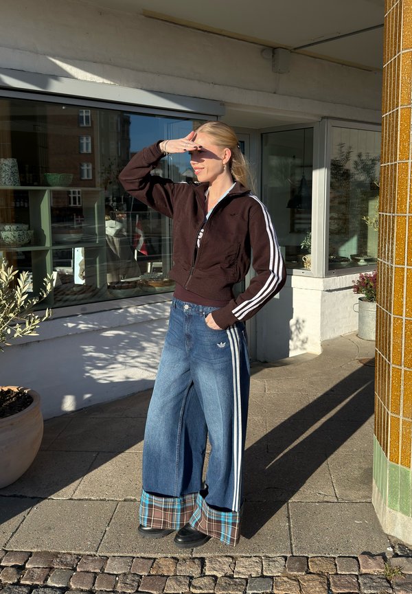 Jonge vrouw die haar ogen afschermt tegen het zonlicht, gekleed in een donker vest met ritssluiting en wijde jeans met geruite boorden, staand op een stadsstoep.
