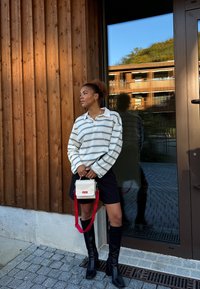 Stripete langermet skjorte i lyse farger, svarte shorts, svarte støvler til knærne, og en liten kremfarget veske med rød stropp. Trebakgrunn.